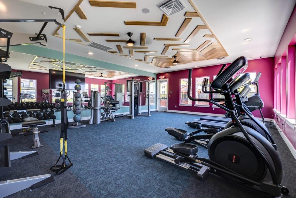 A modern gym in an apartment complex featuring treadmills, ellipticals, a weight rack, and various exercise equipment in a room with a colorful design and patterned ceiling.