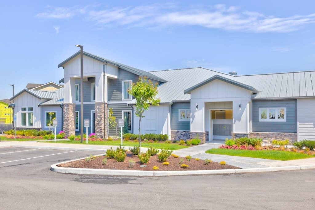 A modern apartment complex with a light gray and white exterior, featuring a metal roof and stone accents. The building is surrounded by landscaped gardens with shrubs and a small tree, located near a paved parking area under a clear blue sky.
