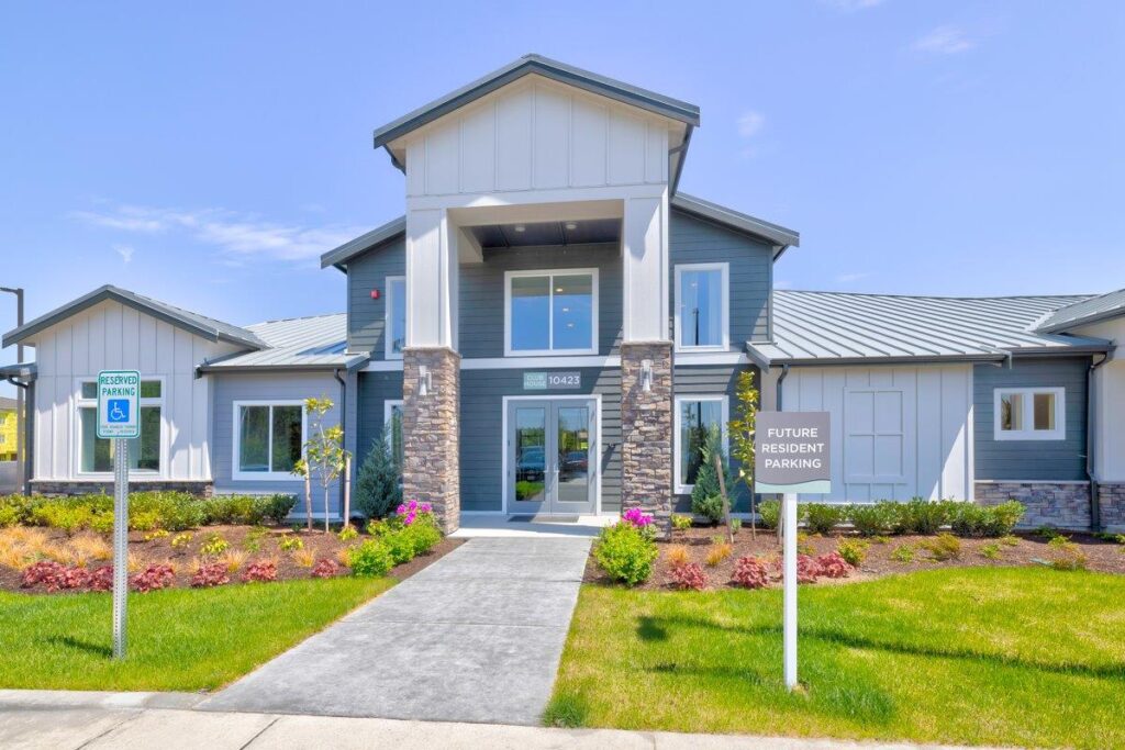 A modern apartment complex entrance with a pathway leading to a building featuring light gray siding and stone accents, surrounded by well-maintained landscaping and signs for reserved and future resident parking.