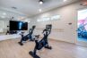 A gym room in an apartment complex featuring two stationary bikes with digital screens, a large wall mirror, and light-colored wooden flooring.