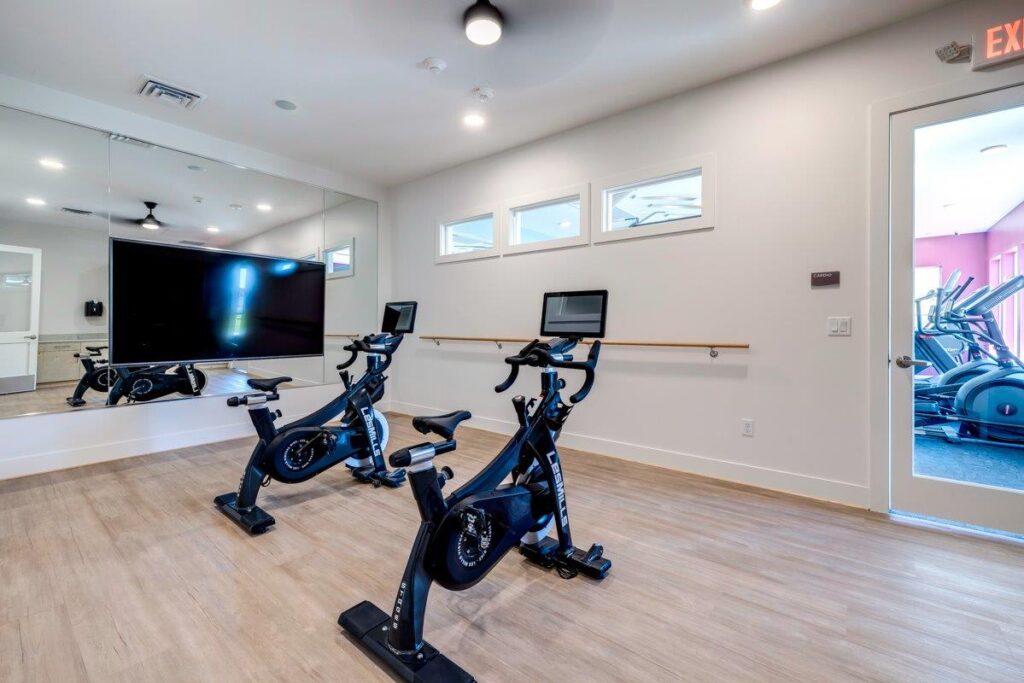 A gym room in an apartment complex featuring two stationary bikes with digital screens, a large wall mirror, and light-colored wooden flooring.