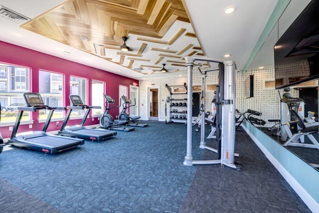 A well-equipped gym in an apartment complex with treadmills, ellipticals, weight machines, and free weights. The room features a unique wooden ceiling design, large mirrors, and a vibrant pink accent wall with windows overlooking the exterior.