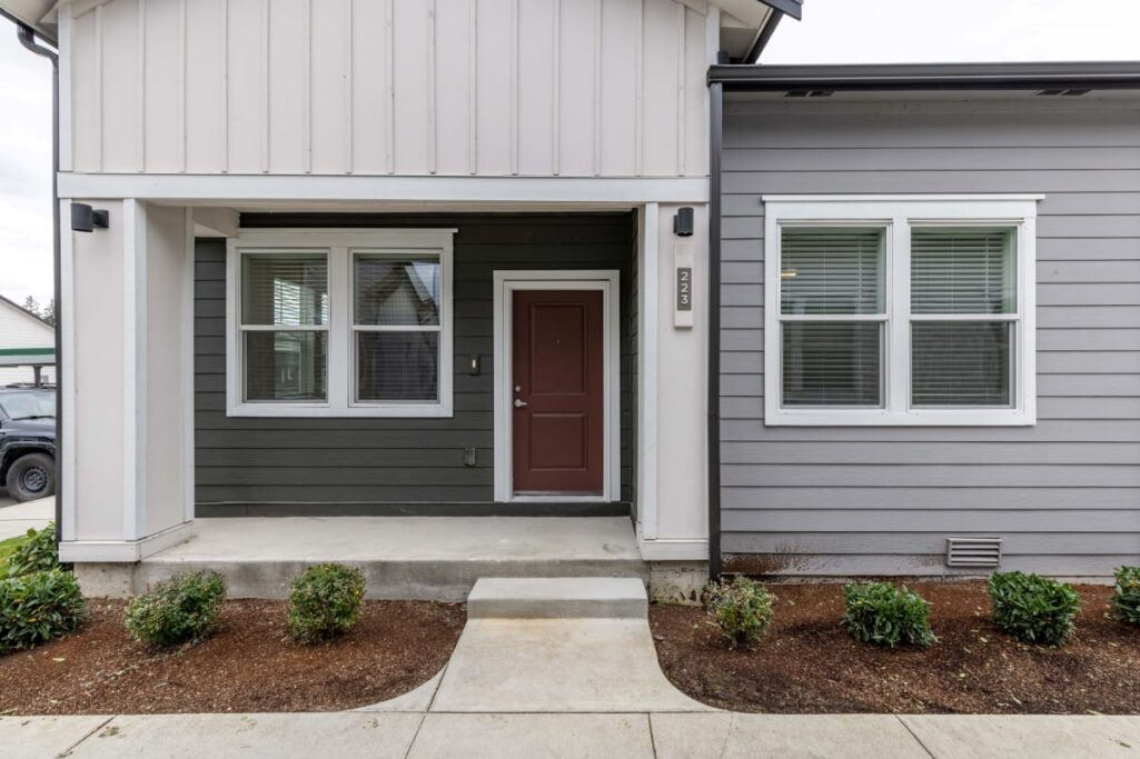 Entrance of a modern apartment with a brown door and gray siding, surrounded by small shrubs.