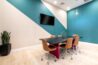Small conference room with a long black table, brown chairs, teal accent wall, and a wall-mounted TV.