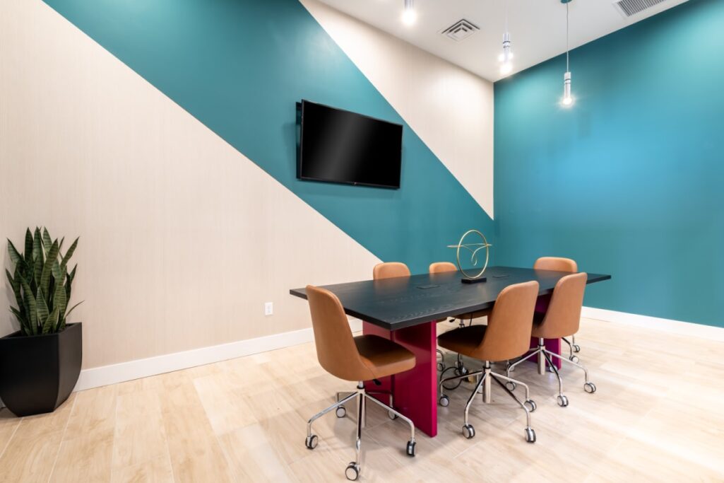 Small conference room with a long black table, brown chairs, teal accent wall, and a wall-mounted TV.