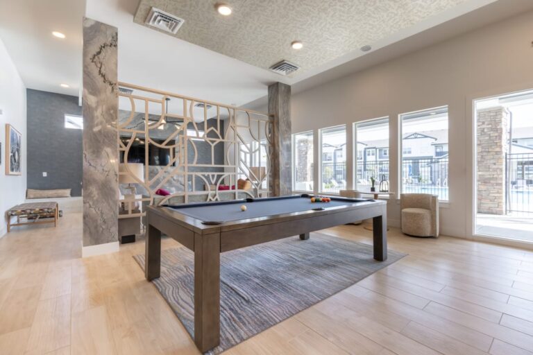A modern apartment complex lounge with a pool table in the center, stylish seating, and large windows offering a view of the exterior patio area.