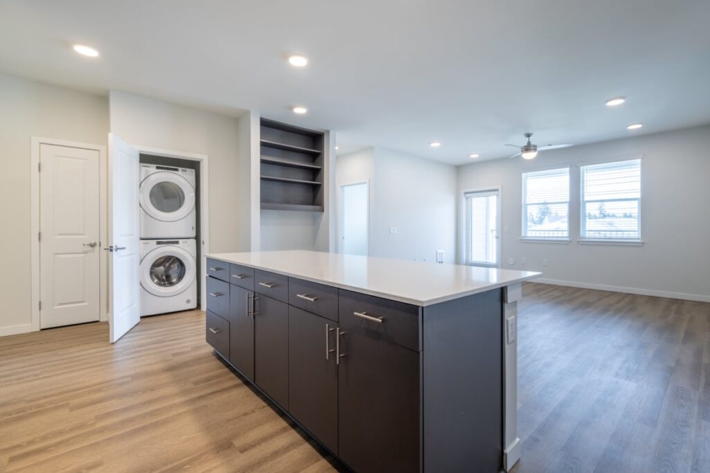 Modern apartment interior featuring a kitchen with a large island, cabinets, and a view of the living area with a washing machine visible in an open closet.