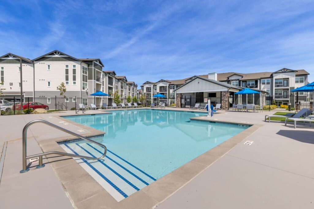 A modern apartment complex featuring multiple buildings with a central outdoor swimming pool, surrounded by lounge chairs and umbrellas, under a clear blue sky.
