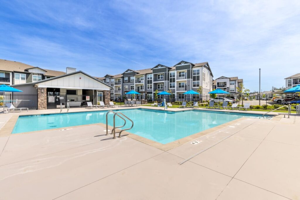 A modern apartment complex with a large outdoor swimming pool surrounded by lounge chairs and umbrellas under a clear blue sky.