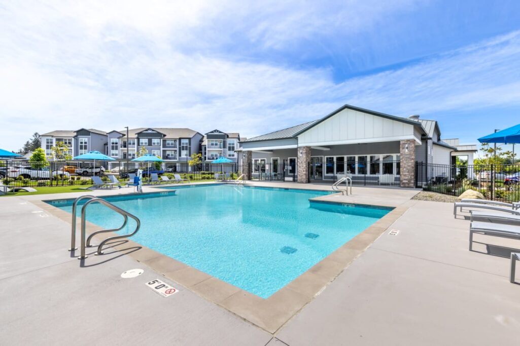 Outdoor swimming pool at Solace Rainer Ridge, surrounded by lounge chairs and blue umbrellas, with multi-story residential buildings in the background.