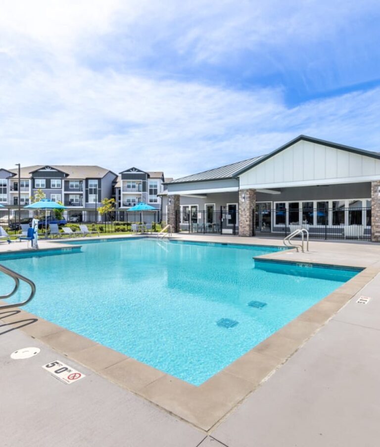 Outdoor swimming pool at Solace Rainer Ridge, surrounded by lounge chairs and blue umbrellas, with multi-story residential buildings in the background.