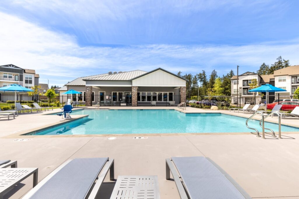 Outdoor pool area of an apartment complex surrounded by lounge chairs and blue umbrellas, with a clubhouse in the background under a partly cloudy sky.