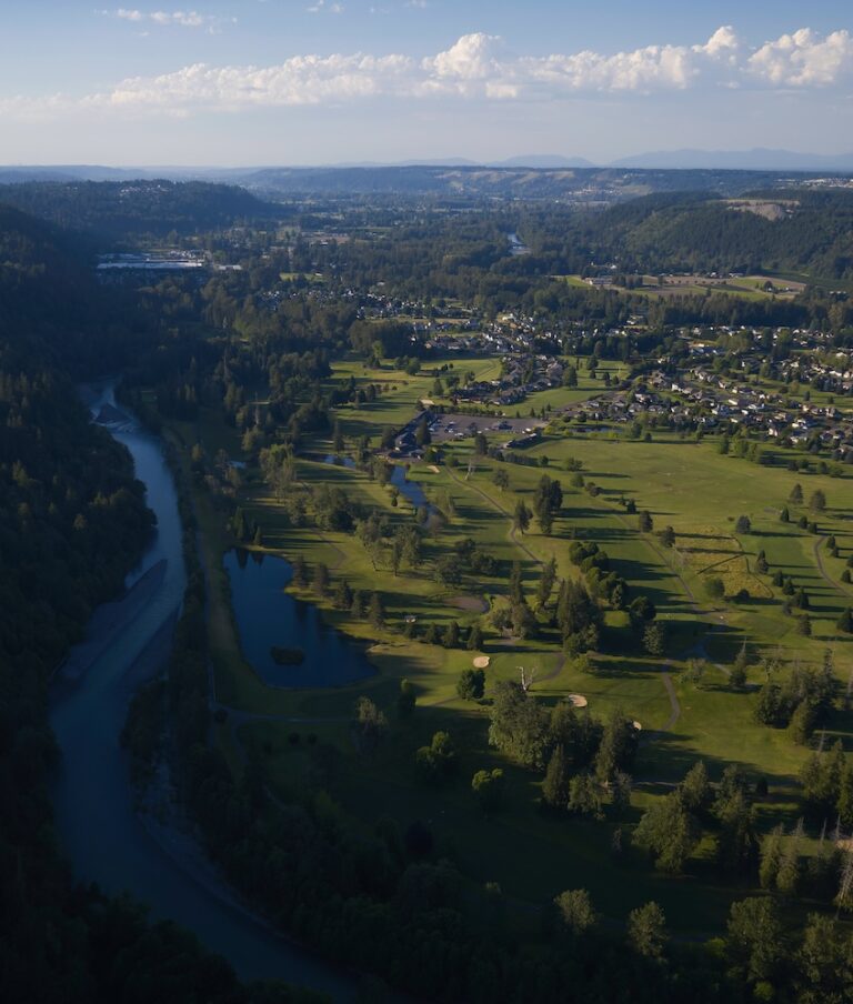 Aerial view of a winding river surrounded by lush forested hills on the left and a green golf course with adjacent residential areas on the right.