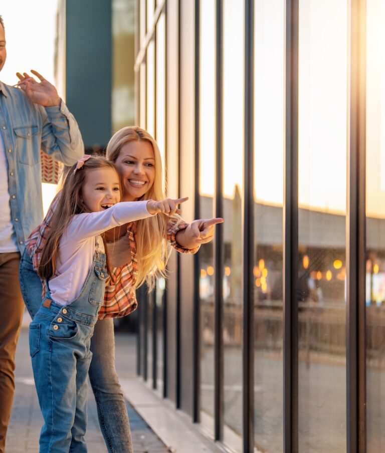 A smiling family of three, with the father carrying shopping bags, the mother and daughter pointing at something through a storefront window during sunset.