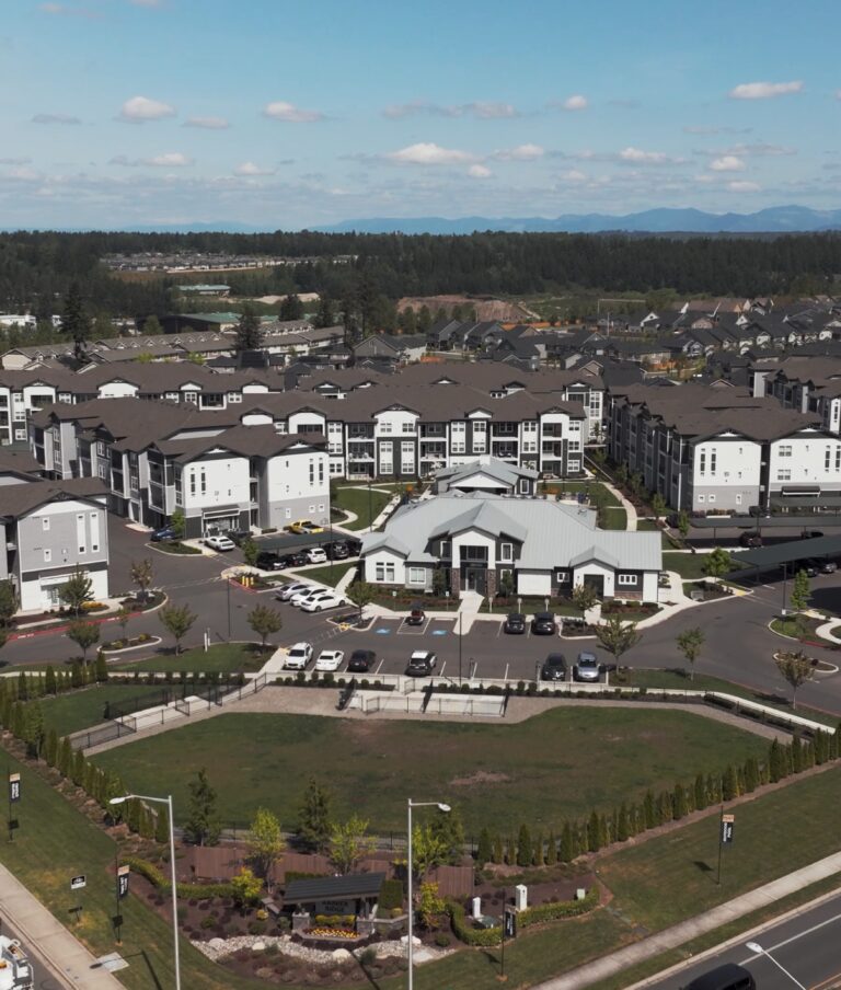 Aerial view of a large residential complex with multiple multi-story buildings surrounded by roads, greenery, and parking areas. The complex is set amidst a suburban environment with open fields and trees in the background.